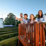 A family of four stands on a wooden platform overlooking a hedge maze, with a white windmill and trees in the background under a clear sky.