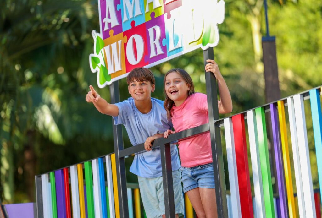 Two children stand behind a colorful fence, smiling and pointing forward under a sign that reads "AMAZE WORLD," with greenery in the background.