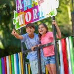Two children stand behind a colorful fence, smiling and pointing forward under a sign that reads "AMAZE WORLD," with greenery in the background.