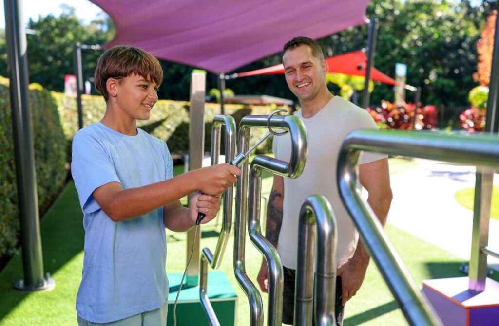 A boy uses a metal skill game outdoors while an adult man watches and smiles. The scene is set in a park with colorful shade structures.