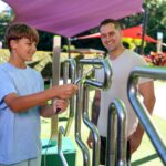 A boy uses a metal skill game outdoors while an adult man watches and smiles. The scene is set in a park with colorful shade structures.