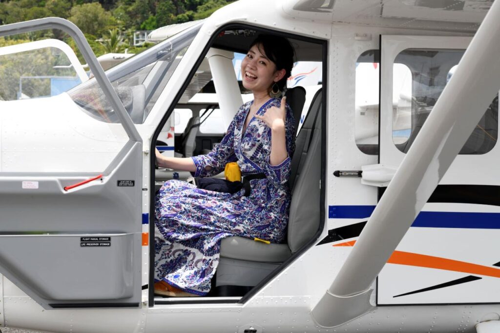A woman in a patterned dress sits in the pilot seat of a small airplane, smiling and giving a thumbs-up with the cockpit door open.