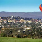 A red and orange hot air balloon floats above a town with suburban houses and buildings, set against distant hills and a clear blue sky.