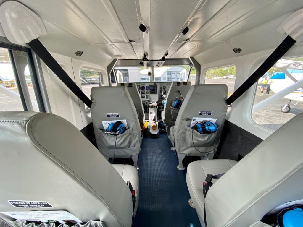 Interior view of a small airplane showing four gray seats, safety harnesses, and the cockpit with flight controls and instrument panel visible at the front.