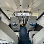 Interior view of a small airplane showing four gray seats, safety harnesses, and the cockpit with flight controls and instrument panel visible at the front.
