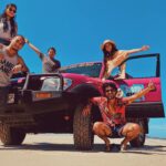Five people pose cheerfully around a pink off-road vehicle parked on a sandy beach under a clear blue sky.