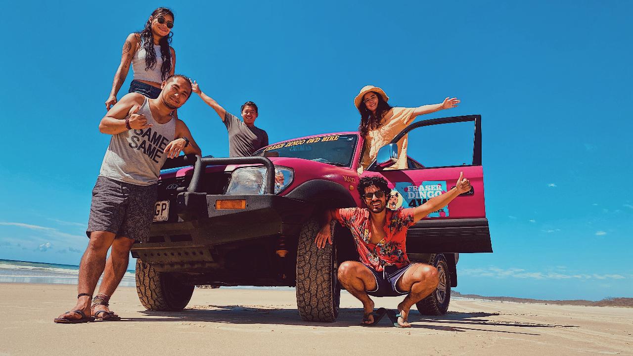 Five people pose cheerfully around a pink off-road vehicle parked on a sandy beach under a clear blue sky.