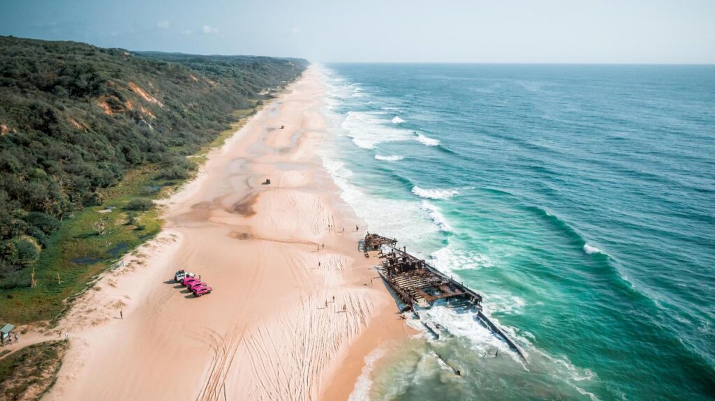 Aerial view of a long sandy beach with a rusted shipwreck near the shoreline and several vehicles parked on the sand beside coastal vegetation.