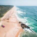 Aerial view of a long sandy beach with a rusted shipwreck near the shoreline and several vehicles parked on the sand beside coastal vegetation.