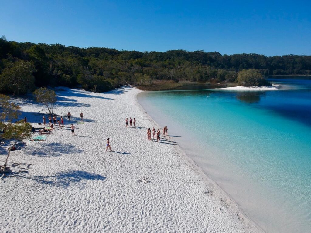 People walk and relax on a white sandy beach next to clear blue water, with trees and vegetation in the background under a clear blue sky.