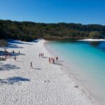 People walk and relax on a white sandy beach next to clear blue water, with trees and vegetation in the background under a clear blue sky.