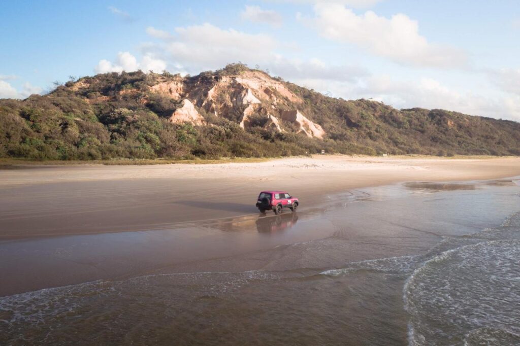 A red SUV drives along a sandy beach with a rocky, tree-covered hill in the background under a partly cloudy sky.