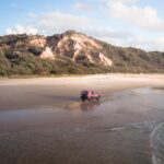 A red SUV drives along a sandy beach with a rocky, tree-covered hill in the background under a partly cloudy sky.