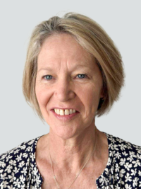 A middle-aged woman with short blonde hair, wearing a dark floral top and a necklace, smiles in front of a plain light grey background.