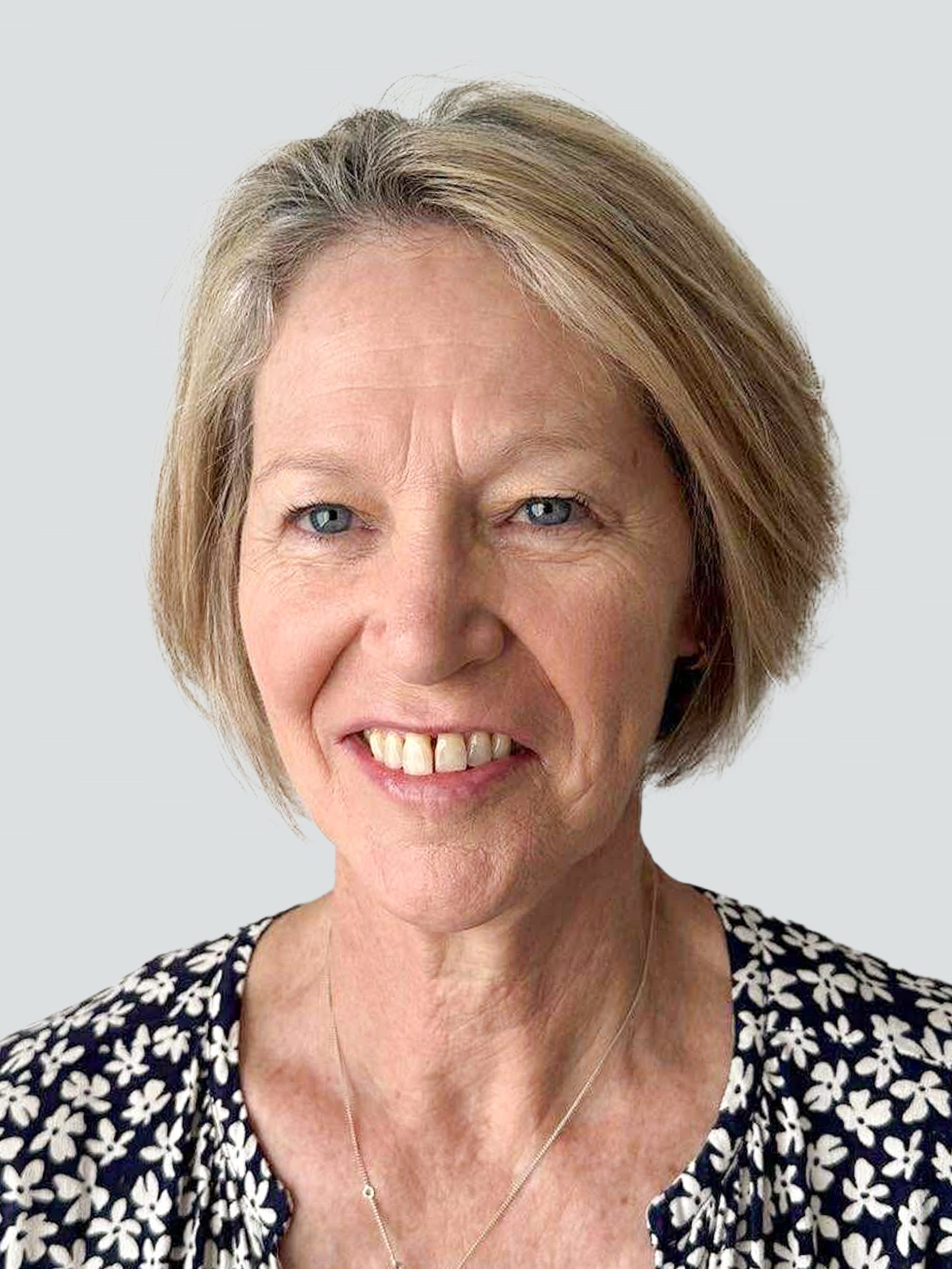 A middle-aged woman with short blonde hair, wearing a dark floral top and a necklace, smiles in front of a plain light grey background.