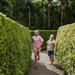 Two children are running along a gravel path between tall, neatly trimmed hedge walls in a garden or hedge maze, with trees in the background.
