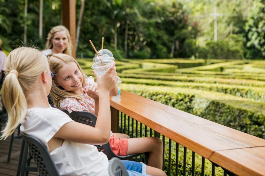 Two young girls sit at an outdoor table overlooking a hedge maze, holding up drinks and smiling, with an adult woman seated behind them in a garden setting.