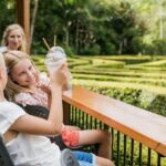 Two young girls sit at an outdoor table overlooking a hedge maze, holding up drinks and smiling, with an adult woman seated behind them in a garden setting.