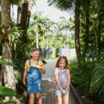 Two young girls walking and smiling on a wooden path surrounded by lush green tropical plants and trees.