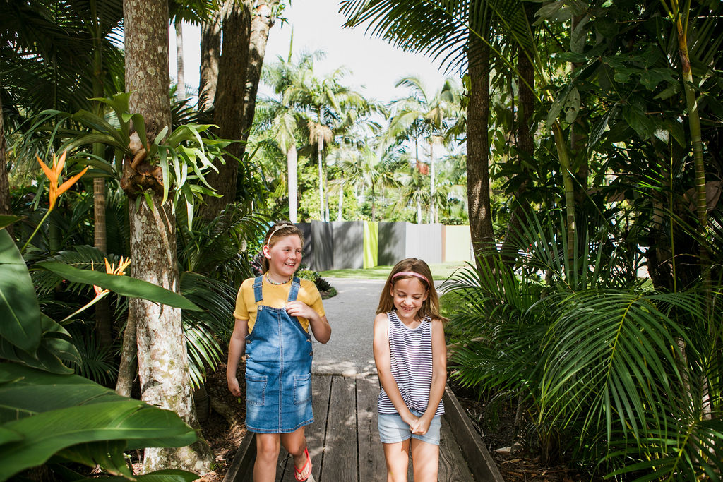 Two young girls walking and smiling on a wooden path surrounded by lush green tropical plants and trees.