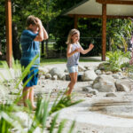 Three children play barefoot near a water fountain in an outdoor park area surrounded by rocks, plants, and shaded structures.