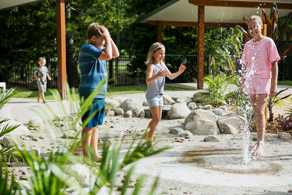 Three children play barefoot near a water fountain in an outdoor park area surrounded by rocks, plants, and shaded structures.