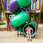 Two children at a playground; one girl slides out of a green and purple tube slide while another girl walks nearby.