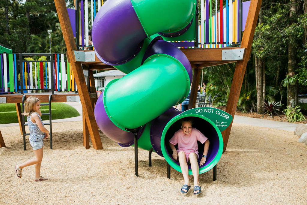 Two children at a playground; one girl slides out of a green and purple tube slide while another girl walks nearby.