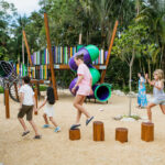 Children playing on wooden stepping posts and climbing equipment in an outdoor playground with colorful slides, surrounded by trees and greenery.