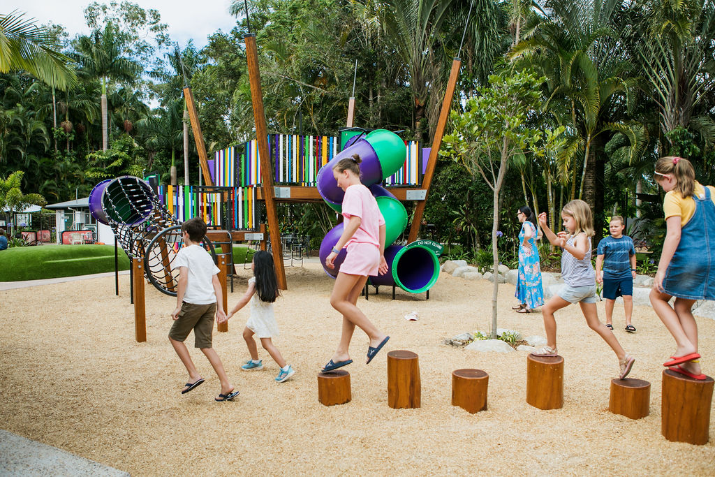 Children playing on wooden stepping posts and climbing equipment in an outdoor playground with colorful slides, surrounded by trees and greenery.