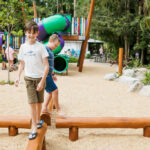 Two boys balance on wooden beams in an outdoor playground, with colorful slides and other children visible in the background.