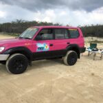 A pink 4WD vehicle is parked on sandy ground near two blue camping chairs and a small table with items, with bushes and cloudy sky in the background.