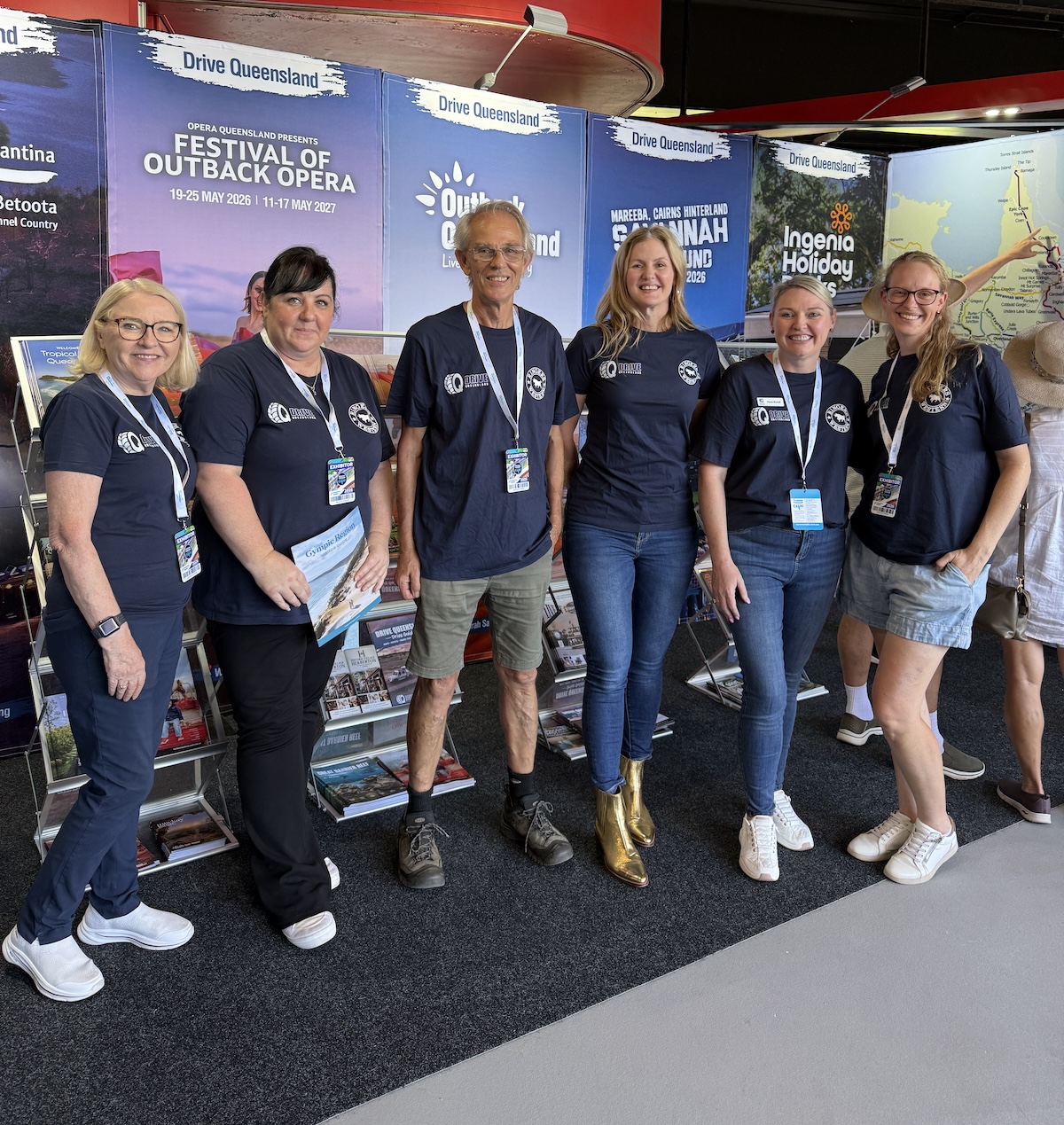 Six people in matching navy shirts stand smiling in front of a tourism booth with Queensland travel posters and brochures on display racks behind them.