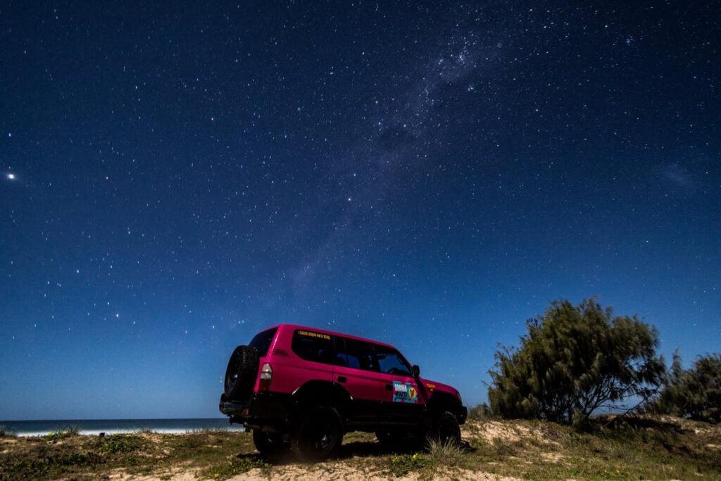 A red off-road vehicle parked on sandy terrain near the ocean under a clear, star-filled night sky.