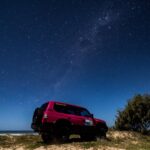 A red off-road vehicle parked on sandy terrain near the ocean under a clear, star-filled night sky.