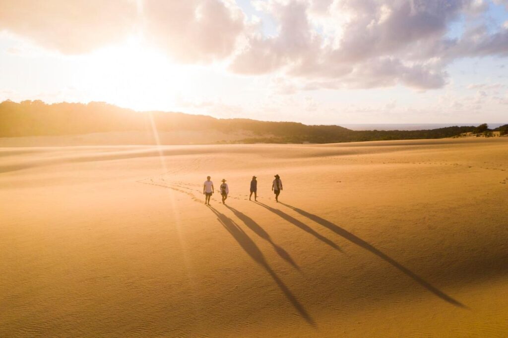 Four people walk across a sunny desert landscape, casting long shadows on the sand, with clouds and hills visible in the background.