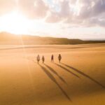 Four people walk across a sunny desert landscape, casting long shadows on the sand, with clouds and hills visible in the background.