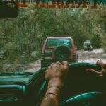 A person drives a car on a dirt road through a forest, following two other vehicles visible ahead.