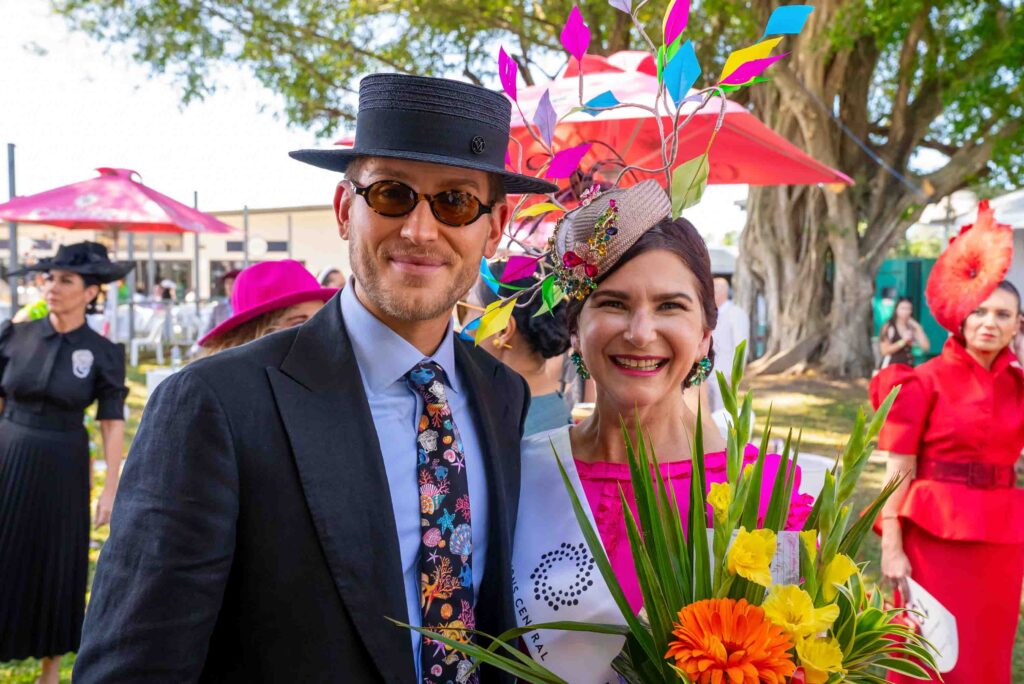 A man in a suit and a woman in a bright floral hat and sash stand together outdoors, smiling and holding a bouquet, with other people and colorful hats in the background.