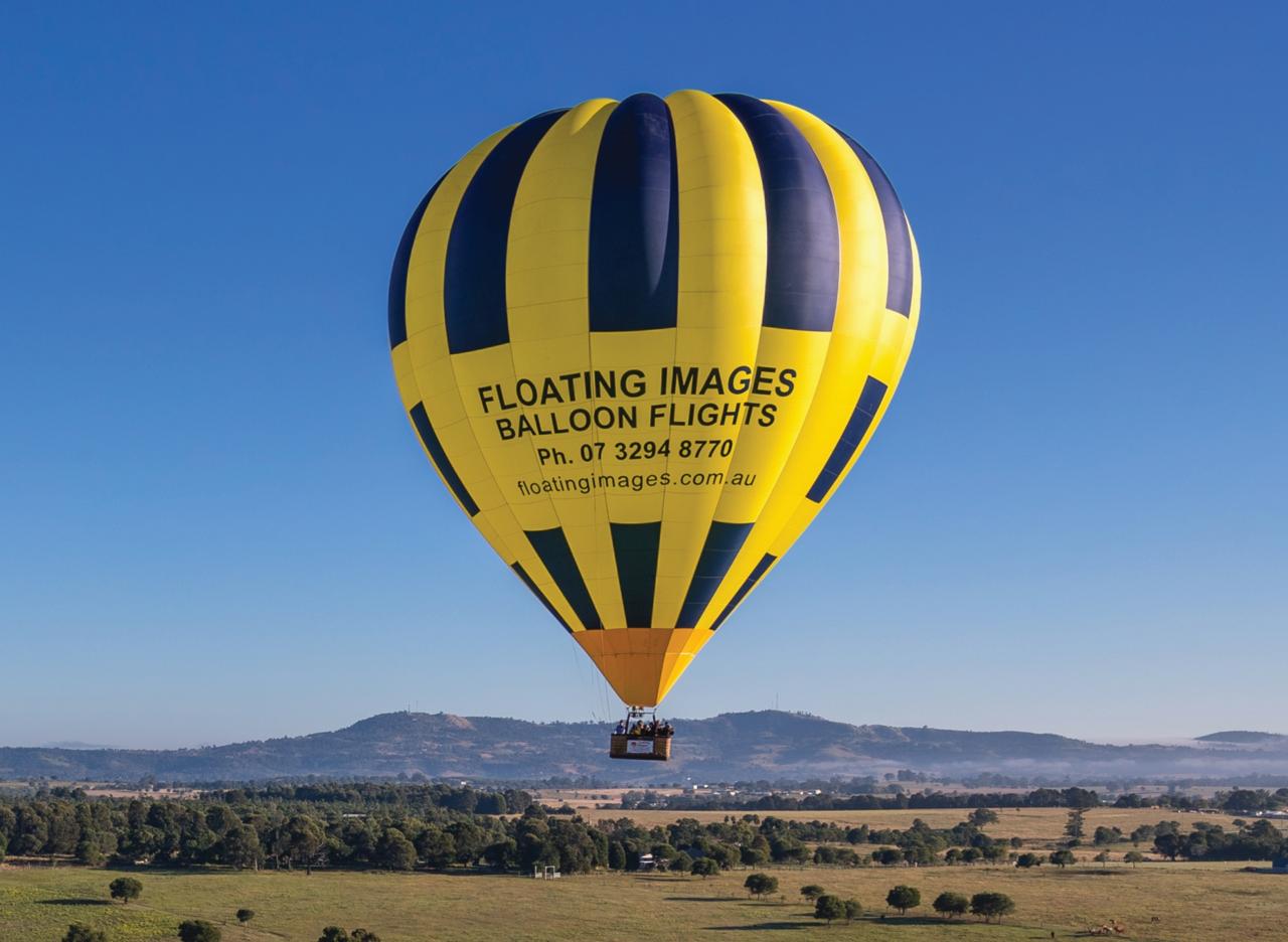 A yellow and blue hot air balloon with “Floating Images Balloon Flights” and a phone number floats above a rural landscape with fields and distant hills.