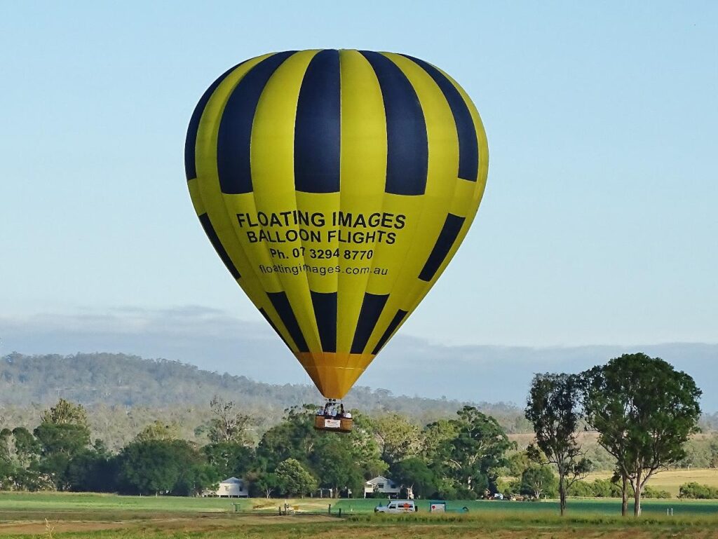 A yellow and black striped hot air balloon with advertising text floats above a grassy field with trees and hills in the background.