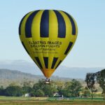 A yellow and black striped hot air balloon with advertising text floats above a grassy field with trees and hills in the background.