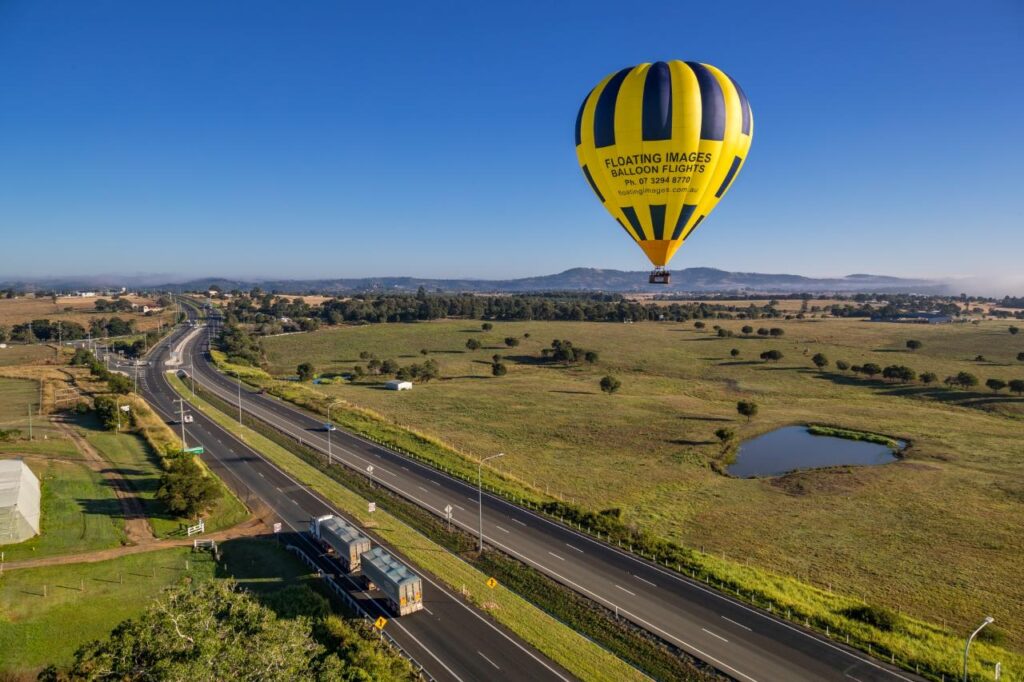 A yellow and black hot air balloon floats above a rural highway and green fields under a clear blue sky.