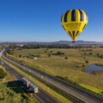 A yellow and black hot air balloon floats above a rural highway and green fields under a clear blue sky.