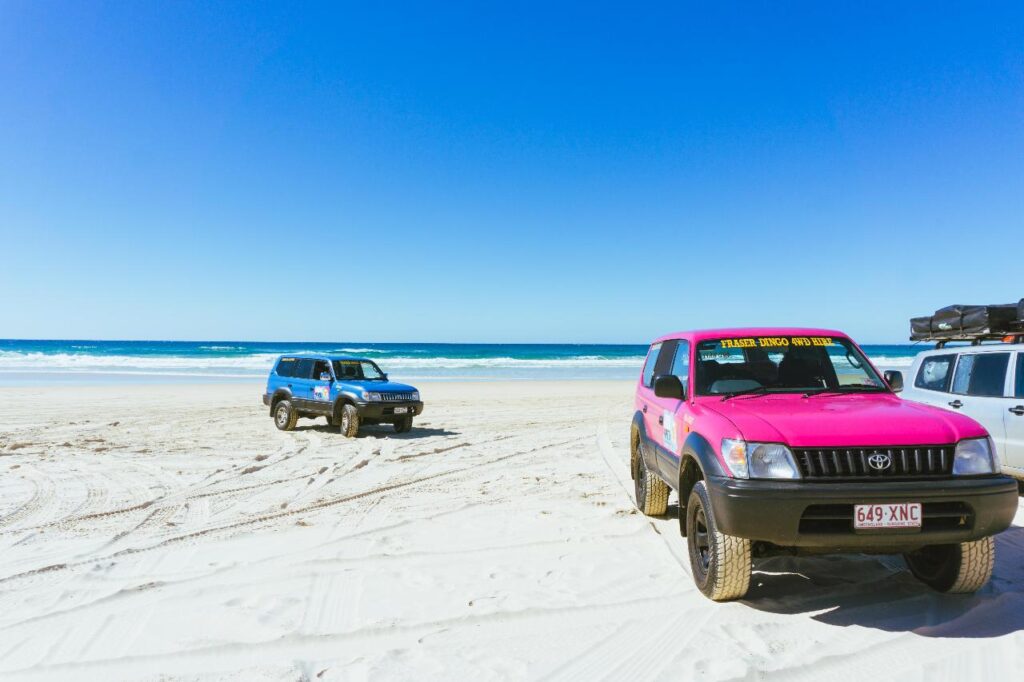 Two SUVs, one blue and one pink, are parked on a sandy beach under a clear blue sky, with the ocean visible in the background.