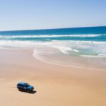 A blue SUV drives along an empty sandy beach near the shoreline with ocean waves under a clear blue sky.