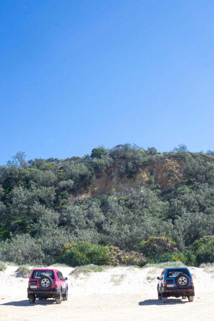 Two off-road vehicles are parked on sandy ground in front of a green hillside under a clear blue sky.