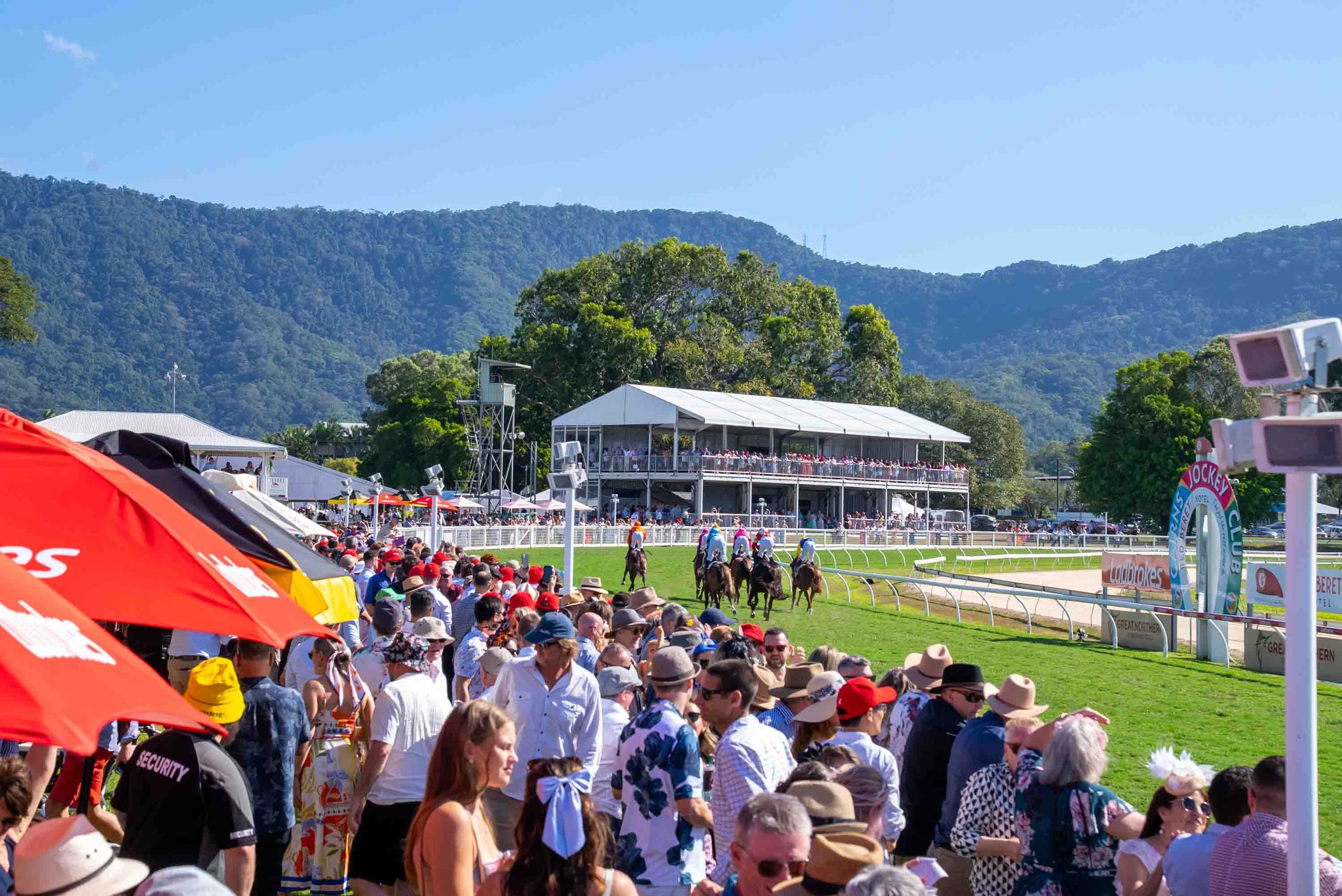 A large crowd watches horses and jockeys prepare for a race at an outdoor racetrack on a sunny day, with mountains and grandstands in the background.