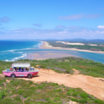 A pink amphibious tour vehicle drives along a coastal dirt path with a scenic view of the beach, ocean, sand dunes, and greenery under a clear blue sky.