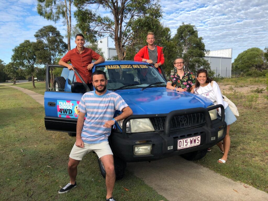 Five people pose in front of a blue 4WD vehicle parked on a paved path with trees and a building in the background.
